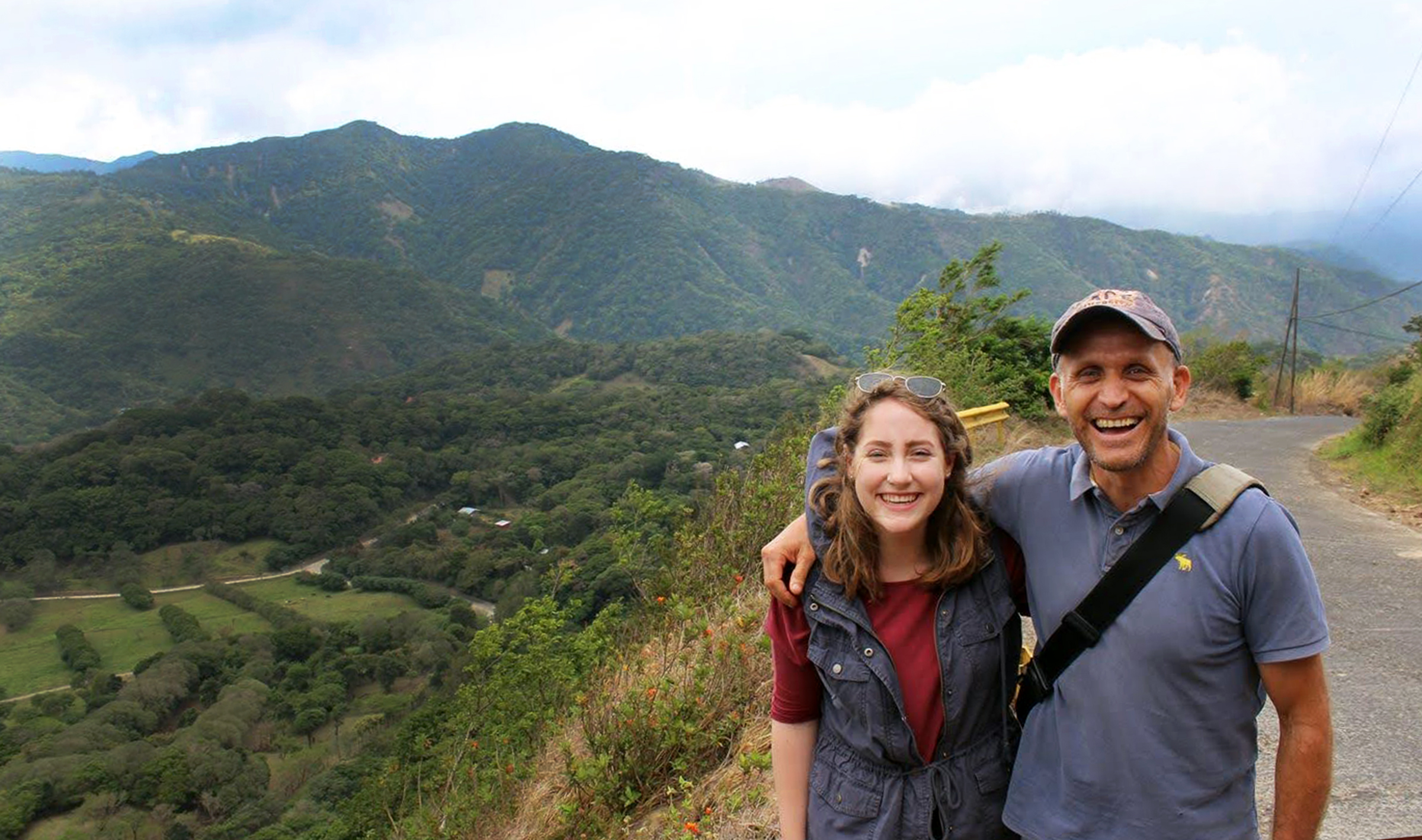 Image shows two people standing arm in arm smiling in front of a green mountain